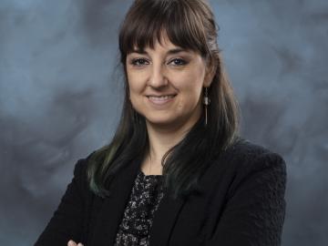 Elena Tajuelo Rodriguez is standing with her arms crossed posing for a photo in front of a blue and dark navy background