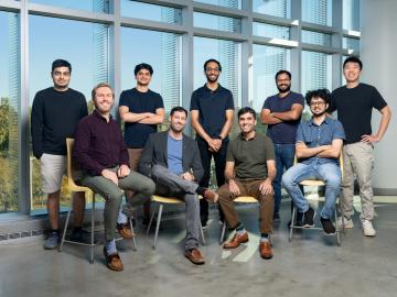 Nine men are pictured here standing in front of a window, posing for a group photo with 5 standing and 4 sitting. 