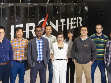 Pictured here are 9 scientists standing in a line in front of the frontier supercomputer logo/computer