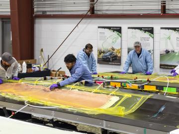 5 scientists in blue and white coats are leaning over the wind blades covered in orange and yellow material