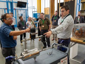 Two scientists are standing in the lab pointing at an object on the table, both wearing glasses. 