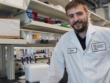 Drew Marquardt in a lab coat leaning on a counter in front of his work station.