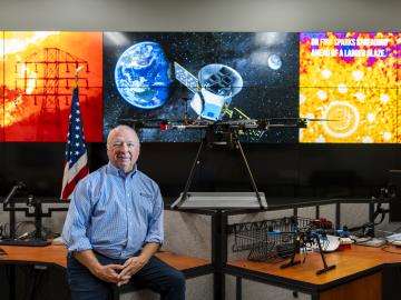 ORNL researcher is sitting on a desk with his hands crossed, three screens behind him depicting work in satellites and space. Pictures on the left and right are orange in color while the middle photo is blue and reflects an image from space