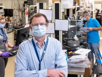 Hugh O'Neill standing at the head of a lab space wearing a mask while other masked scientists work behind him.