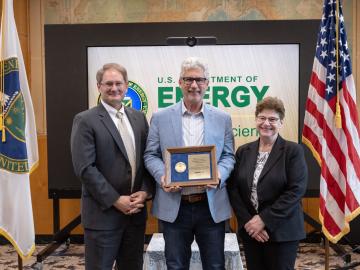 three well-dressed, smiling people stand between a flag with the U.S. Department of Energy seal and the American flag. The person in the center holds an award plaque