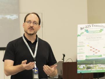 A male researcher is standing next to a poster board displayed on an easel to the right. The researcher is dressed in professional attire, and the poster board is positioned beside him, showing research or visual information.