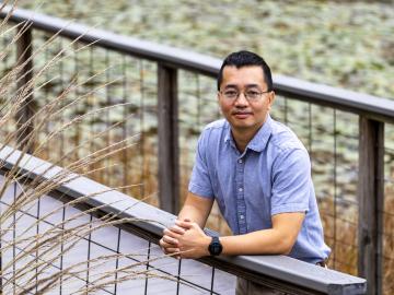 ORNL researcher Phong Le poses for a photo outside on a walkway bridge over the pond. The photo is framed with brown and green plants