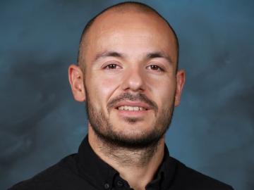 ORNL R&D data scientist Max Pasini is posing for a portrait with a blue background, black button up long sleeve shirt