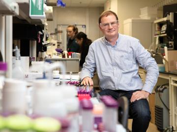 Hugh O'Neil, director or ORNL's Center for Structural Molecular Biology is sitting in the lab on a stool, hand on desk with glasses on. There are lab related items blurred in the foreground.