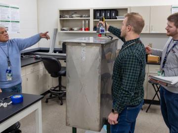 Three researchers are in a lab pointing to a square machine in the middle of the lab. 