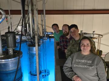 Four researchers are standing next to a research rector that is glowing blue 