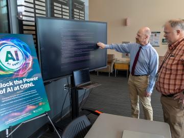 Two ORNL researchers are standing to the right of a computer screen and a poster promoting the AI Expo