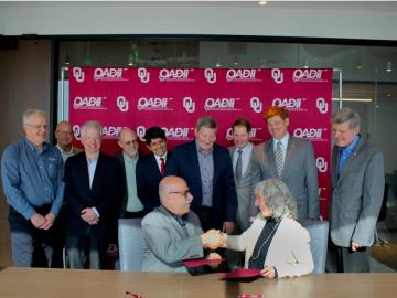 Group of 11 people, 9 standing and two sitting are posing for a photo in front of University of Oklahoma red and white backdrop with UO logo. The two in front are shaking hands