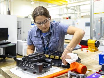 Female MDF researcher leans over component placed on table. Duct work and machinery visible in background.