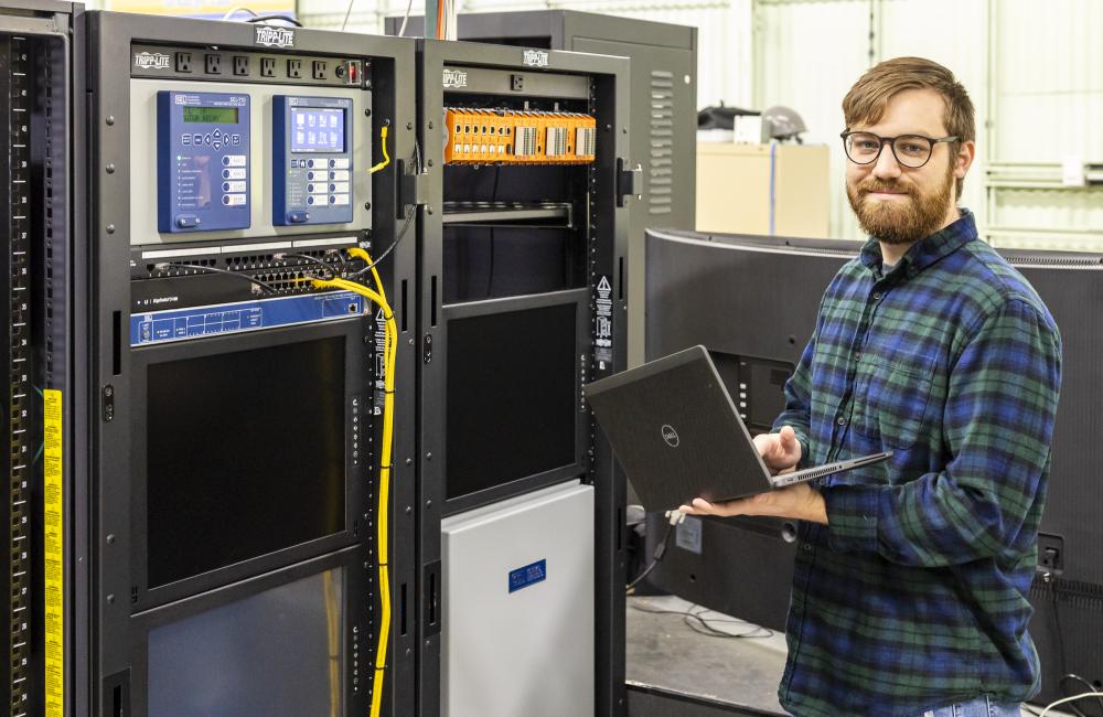 A man wearing glasses and holding a laptop stands near cyber-physical equipment.