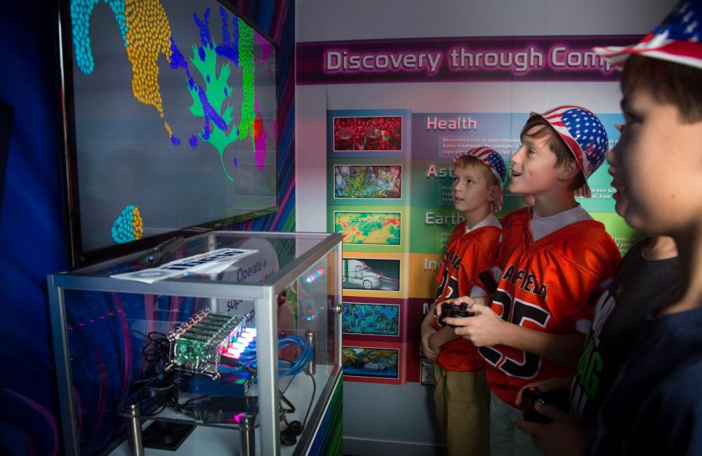 Three boys stand in front of a monitor showing a simulation run by Tiny Titan. 