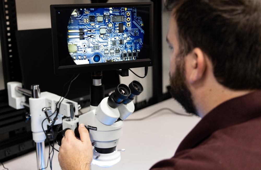 A man looking away from the camera into a microscope with a circuit board