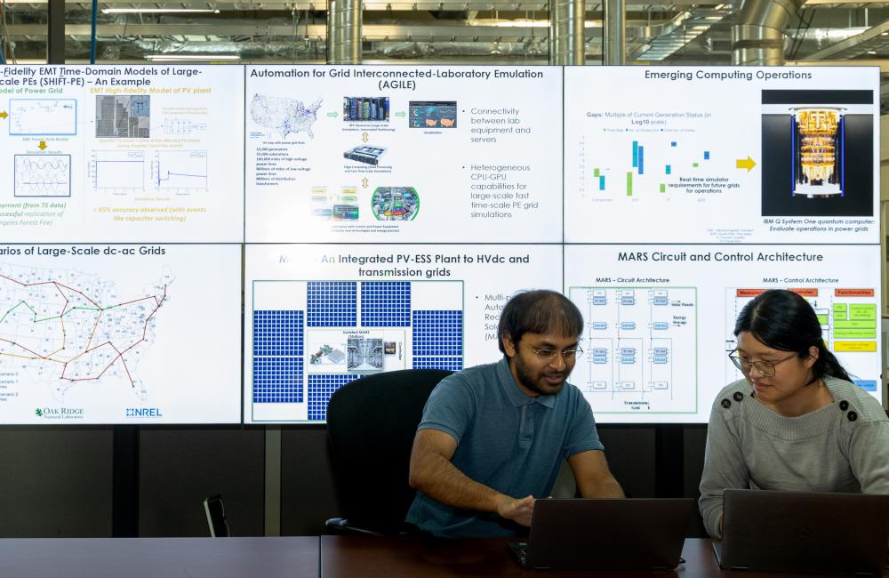 A man and woman researcher look at information on their laptops with a large screen behind them showing research slides