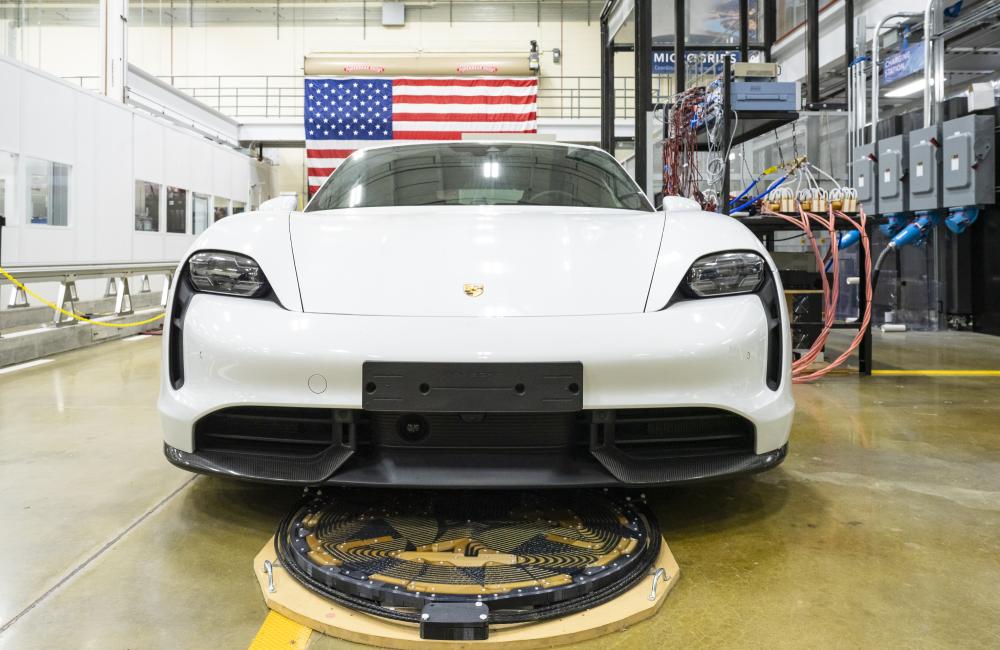 White sportscar parked almost over a wireless charging node of coils ont eh ground, wires and an American flag in background