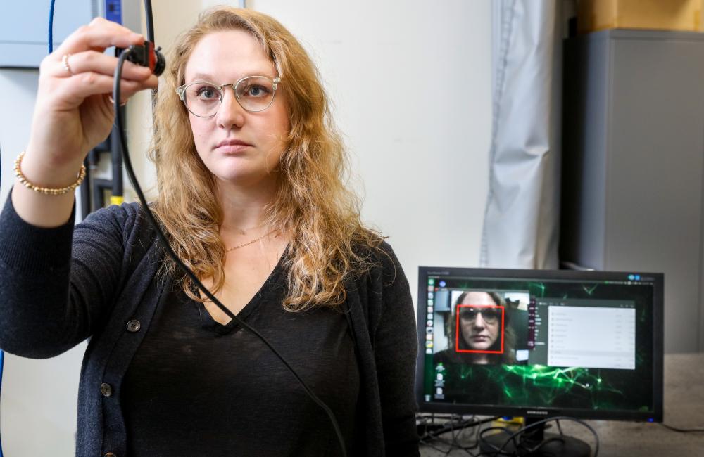 A woman wearing glasses holds a small camera to her face while a computer screen behind her shows a facial recognition software
