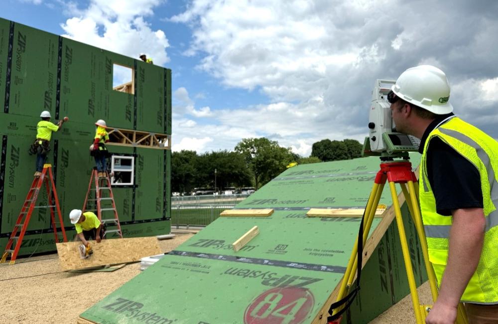Man in bright yellow safety vest and hard hat is looking through a small machine that is pointed at a house being constructed. 