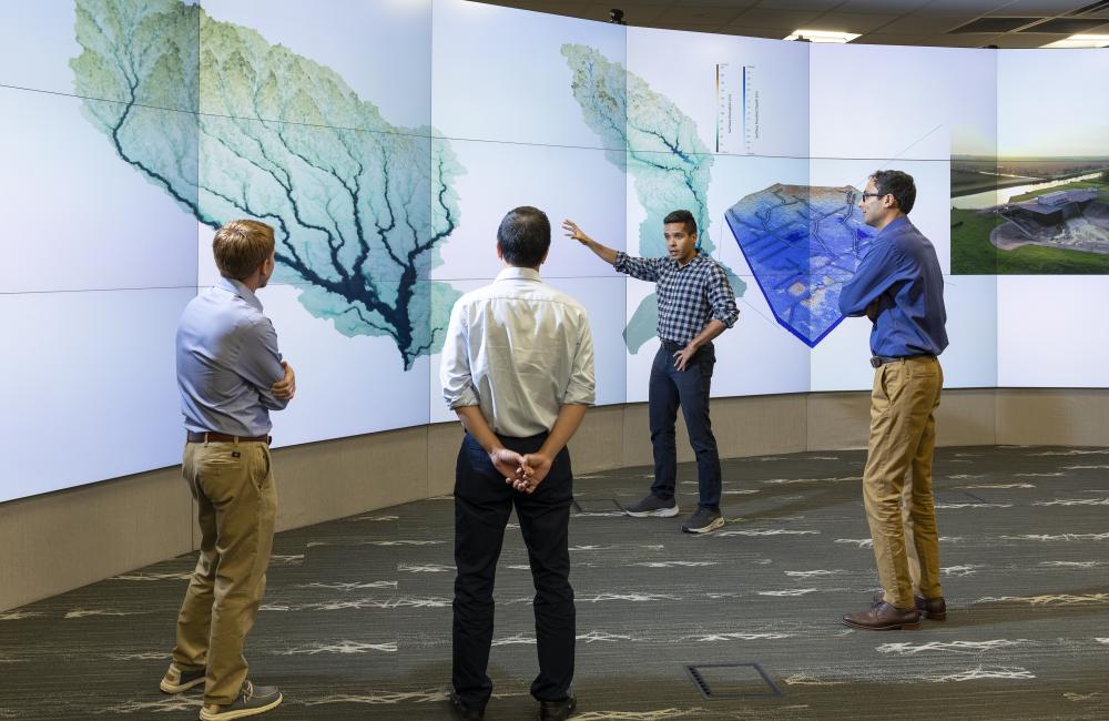 This photo is of four men standing in front of a wall of monitors that are showing a tree looking image.