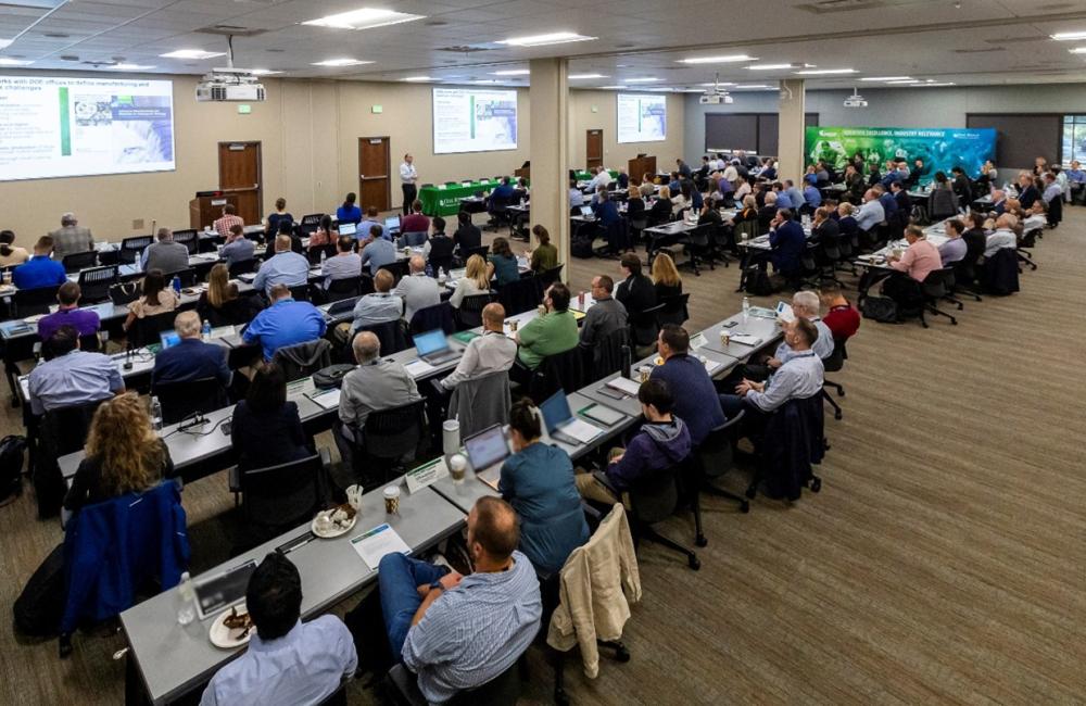 Large group of visitors are listening to a presentation in a large room with three video screens and 10 long desks