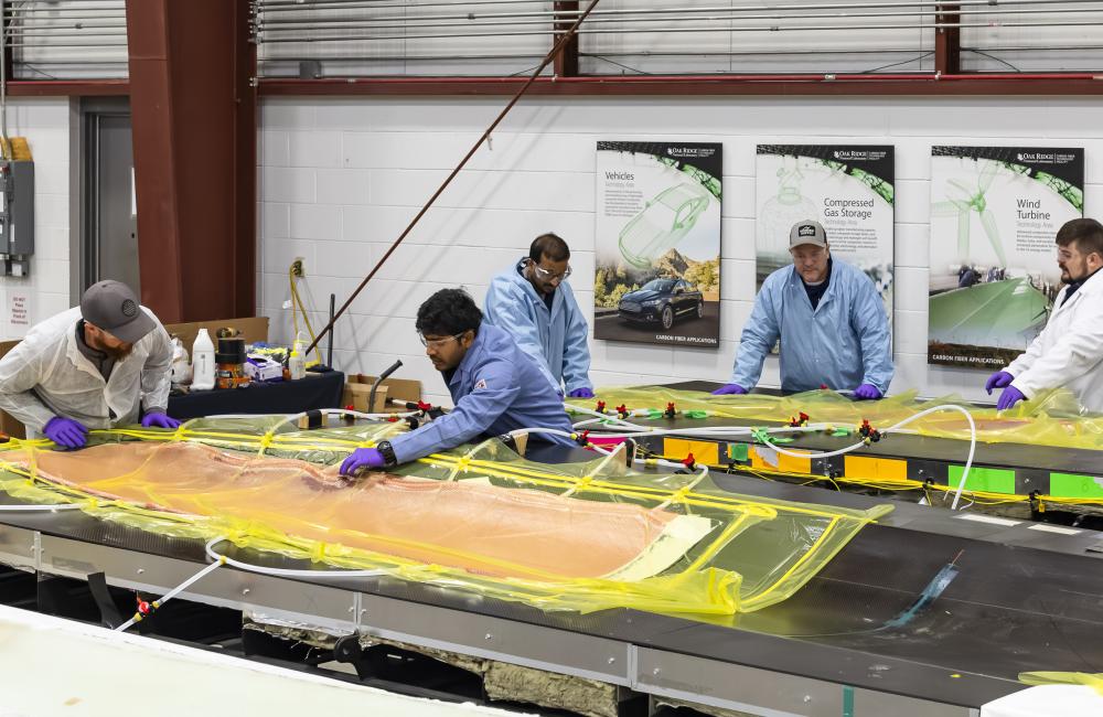 5 scientists in blue and white coats are leaning over the wind blades covered in orange and yellow material