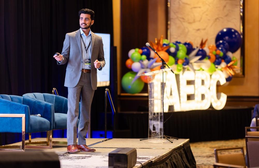 Scientist in a grey suit is standing on stage giving a presentation. There is a TAEBC glowing sign to his right, 
