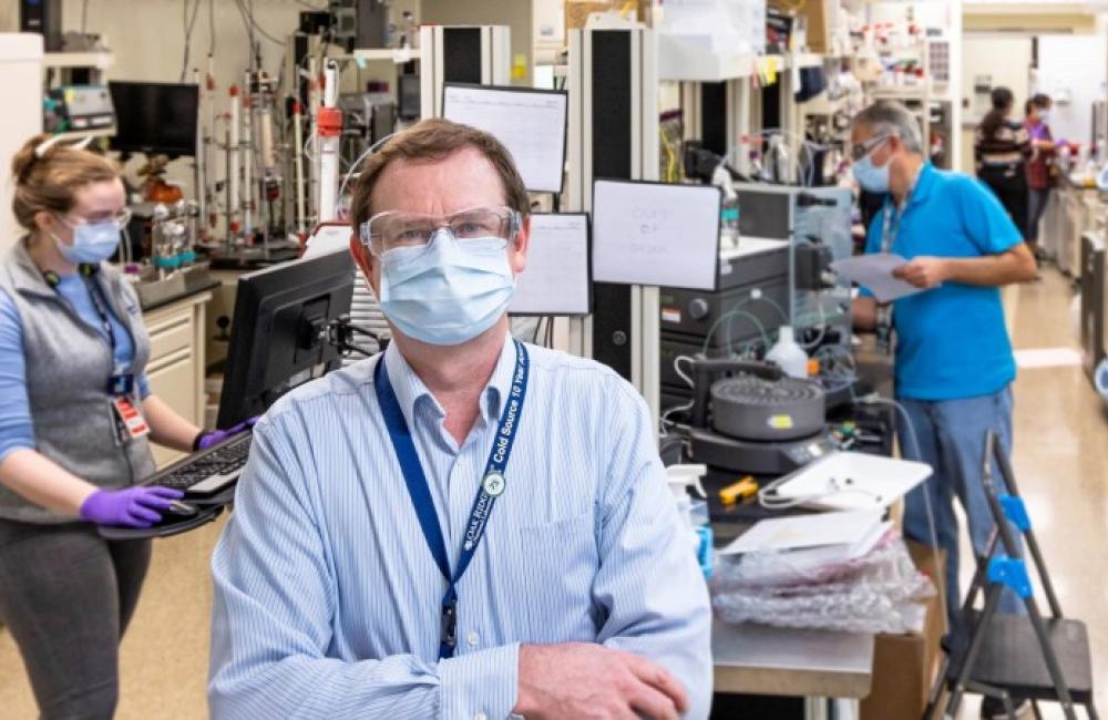Hugh O'Neill standing at the head of a lab space wearing a mask while other masked scientists work behind him.