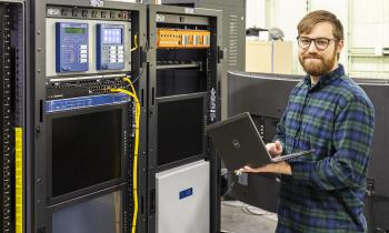 A man wearing glasses and holding a laptop stands near cyber-physical equipment.
