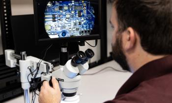 A man looking away from the camera into a microscope with a circuit board