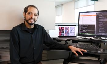 Man in black shirt stands in front of computer screens with maps