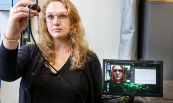 A woman looks into a handheld camera while a computer monitor behind her displays a facial recognition algorithm