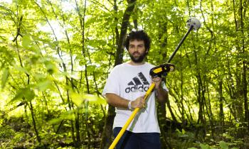 person in a white graphic t-shirt and jeans holding environmental monitoring equipment in a forested area on a sunny day