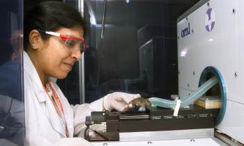 researcher in safety glasses, a white lab coat, and white latex gloves places a small mouse in an imaging machine