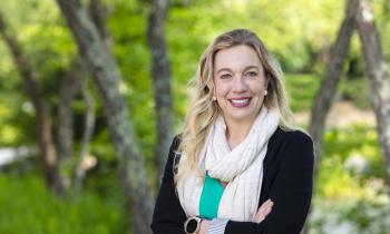 Jessica White-Horton, a female wearing a black sweater and white scarf, poses for a portrait outside in front of green trees.