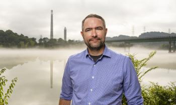 A man stands in front of a lake with steam stacks in the background