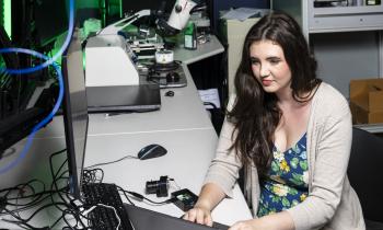 A woman types on a computer with microscopes and computer equipment in the background