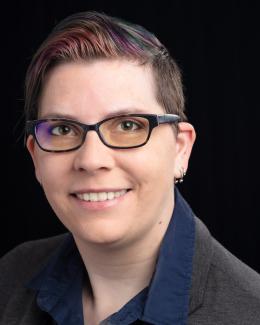 Portrait of Andy Berres. Andy is in their 30s, caucasian with short hair which is brown on the sides and dyed purple and green on top (color subject to change). They are wearing glasses and a navy blue button-up, and a gray jacket and smiling at the camera.