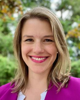 a picture of a blonde woman wearing a white shirt and purple blazer, smiling in front of trees