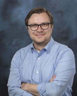 A man in a blue shirt and glasses crosses his arms and smiles for the camera