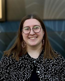 A. Louise Ferris outside, smiling. she has brown glasses on, and is wearing a black and white speckled top. 
