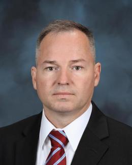 Portrait of a man wearing a white shirt, black coat, and maroon tie with blue diagonal stripes