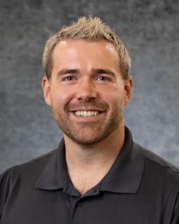 The image shows Liam White smiling with short, light-colored hair, wearing a black collared shirt with the Oak Ridge National Laboratory (Manufacturing Demonstration Facility) logo embroidered on it. The background is a neutral gray gradient.