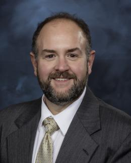 A portrait of a man in a grey suit with a beard on a blue background