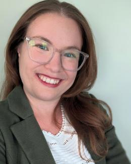 Professional portrait of Ashley, a smiling woman with long auburn hair and clear-framed glasses, wearing a green blazer over a white blouse. She is posed confidently with arms crossed against a soft green background.