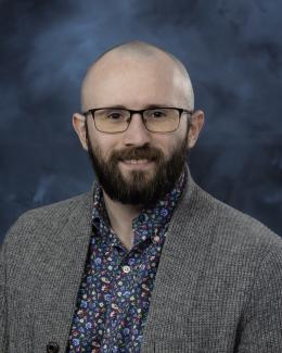 Photo of a bearded man wearing glasses, a floral print shirt, and an unbutton gray cardigan. He is smiling at the camera.