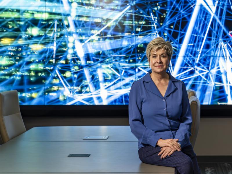 Gina Tourassi is sitting on a table in a conference room in front of a bright screen with blue and white lines