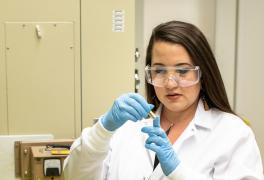 A woman in a lab coat and safety glasses examines a material sample.